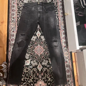 Svarta Lee Luke jeans med slitningar W31 L32 - Säljer ett par svarta jeans från Lee i modellen Luke. Jeansen har snygga slitningar och patchade detaljer på båda benen, femficksmodell och klassisk Lee-logga bak. Materialet är jeans och passformen är slim med raka ben.