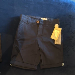 Mörkblå shorts Hampton Republic 140 - Snygga mörkblå shorts från Hampton Republic i klassisk chino-stil. De har uppvikta benslut, bälteshällor, dragkedja och knapp framtill samt två bakfickor med knappar. Perfekta för sommardagar och stilrena outfits.