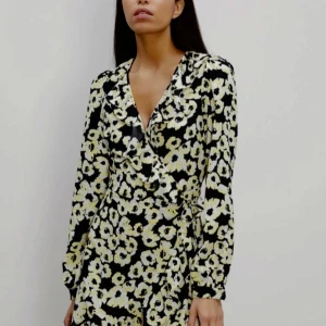 Blommig omlottklänning från Bik Bok - Superfin omlottklänning från Bik Bok med blommigt mönster i svart, vit och gul. Klänningen har långa ärmar, volangdetaljer vid v-ringningen och knyt i midjan. Tillverkad i återvunnen polyester, perfekt för dig som gillar färg och mönster.