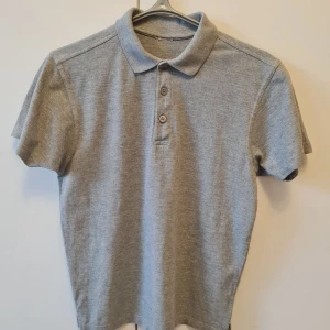 Pikétröja med krage och knappar strl S - Säljer en klassisk grå pikétröja med krage och tre knappar framtill. Tröjan har korta ärmar och är tillverkad i ett mjukt material. Ettikiten är klippt. Storlek S. Kan hämtas i Vallentuna.