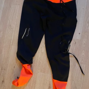 Svarta och orange täckbyxor från Craft - Säljer ett par svarta täckbyxor från Craft med orange detaljer vid midja och benslut. Byxorna har snörning i midjan och inbyggda fötter för extra skydd. Perfekta för utomhusaktiviteter under kalla dagar.