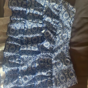 Blåvit volangkjol med mönster - Supersöt kort kjol med blått och vitt mönster och flera volanger. Kjolen har inbyggda shorts på insidan och stängs med dragkedja i sidan. Perfekt för dig som gillar unika prints och vill sticka ut lite extra.