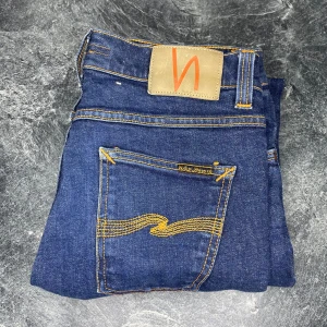 Nudie Jeans blå jeans W30 L32 - Snygga blå jeans från Nudie Jeans med klassiska orange sömmar och logga på bakfickan. Tillverkade i ekologisk bomull och har en rak passform. Perfekta för dig som gillar stilrena och hållbara jeans.