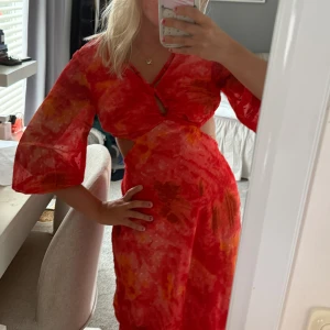 Röd klänning med cut out - Snygg röd och orange  klänning med v-ringning, cut out-detaljer i midjan och vida trekvartsärmar. Klänningen har ett färgstarkt mönster och är tillverkad i ett lätt, flowy material som passar perfekt för sommardagar. Den är använd en gång och i väldigt bra skick 