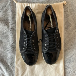Svarta sneakers från Lanvin i mocka - Stilrena svarta sneakers från Lanvin med ovandel i mocka och lackad tå. Skorna har vita gummisulor och klassisk snörning med metallöljetter. Perfekta för dig som gillar en clean och lyxig look med en twist av materialmix.Skorna är  defekterade skriv om mer bilder. Priset kan diskuteras vid snabbt köp. Nypris 4900-5200 Fram på skorna har lite material släppt. Under skorna fins också två små hål. Sulorna har också hål vid hälen men kan bytas ut. Insidan där back på en av skorna finns ett hål.