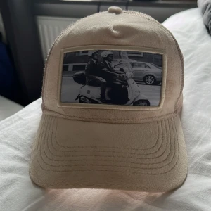 Beige keps från Emilio Pendenza - Snygg beige keps från Emilio Pendenza med mesh på baksidan och en unik svartvit patch framtill med motiv av två personer på scooter. Kepsen har böjd skärm och klassisk trucker-modell. Perfekt för dig som vill sticka ut med en cool accessoar.