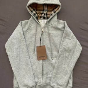 Burberry hoodie - Snygg grå hoodie med dragkedja och klassiskt rutigt foder i huvan. Tröjan har snörning i huvan, två fickor fram och exklusiva detaljer med Burberry-logga på snörändarna. Perfekt för dig som vill ha en stilren och lyxig look. Skriv vid funderingar 
