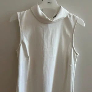 Vit ärmlös polotopp - Snygg vit polotopp med hög krage och ärmlös design. Toppen har en klassisk dragkedja i ryggen och är gjord i ett mjukt, slätt material som känns skönt mot huden. Perfekt att styla med jeans eller kjol för en clean look