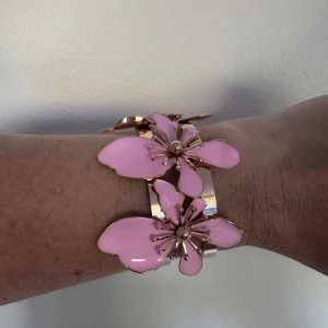 Guldigt armband med rosa blommor - Statement-armband i guldmetall med stora, glansiga blommor i ljusrosa. Blommorna har tydliga detaljer och sitter på en bred, öppen armring. Perfekt för dig som vill sticka ut med en färgglad och lekfull accessoar.