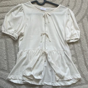 Vit blus med knyt från Gina Tricot - Supersöt vit blus från Gina Tricot med puffiga korta ärmar och tre knyt framtill. Blusen har en lätt volang nertill och är gjord i mjuk bomull. Perfekt för dig som gillar romantisk och trendig stil.