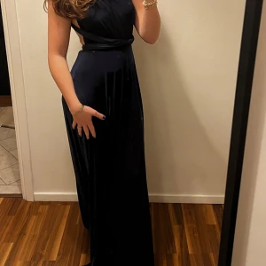 Marinblå långklänning med öppen rygg - Snygg marinblå långklänning med djup v-ringning och markerad midja. Klänningen har en öppen rygg med knytband och är ärmlös. Tyget är glansigt och faller snyggt, perfekt för bal eller fest. Stilren och elegant look som verkligen sticker ut. Köpt för 1200kr på bubbleroom 2024. Storlek 38 man kan göra den minde eller större passar 36-40 storlek.