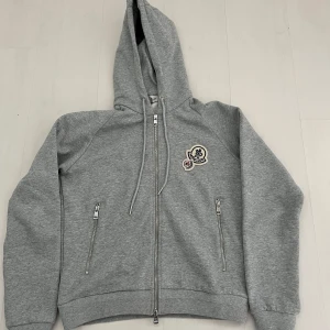 Grå hoodie från Moncler med dragkedja - Snygg grå hoodie från Moncler med huva och två fickor med dragkedja. Hoodin har Moncler-logga på bröstet. ✔️ Fint skick – sparsamt använd.              Skicka gärna ett meddelande vid frågor! 
