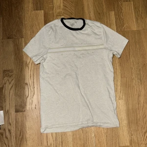 Ljusgrå t-shirt Tommy Hilfiger - Ljusgrå t-shirt från Tommy Hilfiger med broderad logga och diskreta ränder över bröstet. T-shirten har svart kant runt halsen och klassisk passform. Tillverkad i mjuk bomull, perfekt för en clean och stilren look.
