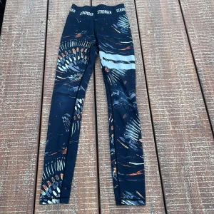 Mönstrade leggings från Stronger XXS - Snygga svarta leggings från Stronger i storlek XXS med coolt abstrakt mönster i vitt, orange och blått. Hög midja med logotyp i midjebandet och två reflekterande ränder på ena benet. Tillverkade i återvunnen polyester och elastan för en stretchig och bekväm passform.                                                       Tyvärr är det ett litet hål vid knät
