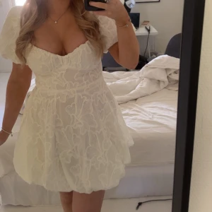 Vit kortklänning med puffärm - Supersöt vit kortklänning med puffärmar och markerad byst. Klänningen har ett stort blommigt mönster i tyget och en lätt voluminös kjol. Använd vid ett tillfälle på min skolavslutning. Den är i super bra skick. Den kostar 450kr ny! 💕💕