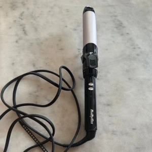 Locktång från BaByliss svart/silver - Locktång från BaByliss i svart och silver med sladd och två knappar för värmereglering. Tången har en klassisk rund form och är perfekt för att skapa snygga lockar eller vågor. Handtaget är svart med vit logga och värmedelen är silverfärgad.