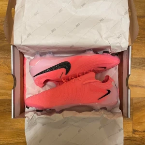 Nike Phantom GX II Elite FG ”Mad Brilliance” - Säljer ett par Nike Phantom GX II Elite FG ”Mad Brilliance”. Slutsålda överallt. Det har en riktigt snygg rosa färg med svarta detaljer och mönstrad Swoosh. Ovandelen är i syntetmaterial med deras kända Gripknit-struktur för extra grepp på bollen. Perfekta för dig som vill sticka ut på planen. Storlek 42, FG dobbar men funkar även på konstgräs. Nypris låg på 3299kr, men jag lägger ut dem för 2500kr. Priset är förhandlingsbart vid en smidig affär. Endast använda en gång.