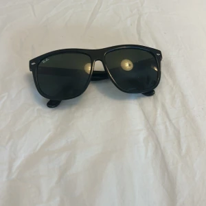 Svarta Ray-Ban Wayfarer solglasögon - Klassiska svarta Ray-Ban Wayfarer solglasögon med mörka glas och stilren design. Bågarna är i blank svart plast och har Ray-Ban-logga på sidan. Perfekta för en cool och tidlös look.