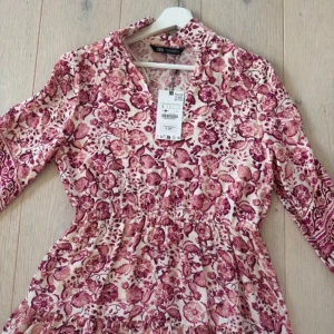 Sommarklänning från Zara - Supersöt helt ny kortklänning från Zara med blommigt mönster i rosa, vinrött och vitt. Klänningen har v-ringning, långa ärmar och markerad midja. Perfekt till sena sommarkvällar eller stranddagar! 