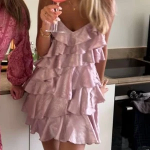 Ljusrosa volangklänning - Supersöt ljusrosa kortklänning från Asos med flera lager volanger och tunna axelband. Klänningen har en glansig finish och v-ringad urringning, perfekt för dig som vill sticka ut. Materialet känns lyxigt och lätt.