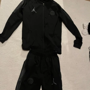 Svart PSG x Jordan tracksuite - Svart tracksuit från Jordan x PSG med dragkedja, långa ärmar och diskreta detaljer på axlarna på tröjan. Tröjan och byxorna har PSG-logga och Jumpman-logga på bröstet. Bra skick