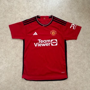 Manchester United hemmatröja 2023/24 - Röd Manchester United fotbollströja från Adidas med svarta detaljer och vita logotyper. Klassisk kortärmad modell med klubbmärke på bröstet och TeamViewer-tryck. Tillverkad i lätt och ventilerande material, perfekt för träning eller match.