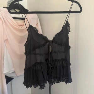 Svart volangtop från Zara - Supersöt svart linnetopp från Zara med tunna axelband och flera lager volanger. Toppen är lätt transparent och har snörning framtill för extra detalj. Perfekt för dig som gillar romantiska och lekfulla vibes.