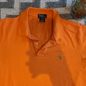 Orange pikétröja från Ralph Lauren - Snygg orange pikétröja från Polo Ralph Lauren i 100% bomull. Pikétröjan är lite blekad vid armhålor, men det syna inte med färgen under solen. Perfekt för dig som vill ha en färgstark och stilren look till sommaren.
