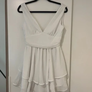 Vit omlottklänning med volang - Supersöt vit kortklänning med omlott och v-ringning både fram och bak. Klänningen har breda axelband och volangig kjol i flera lager som ger en flowy känsla. Perfekt för sommarens alla tillfällen!