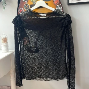 Svart spetsblus med volang från MNG - Säljer en svart, transparent spetsblus från MNG med långa ärmar och volangdetaljer på axlarna. Blusen har en smal passform och är perfekt för dig som gillar en edgy och trendig look. Superfin att styla lager på lager eller med ett linne under.