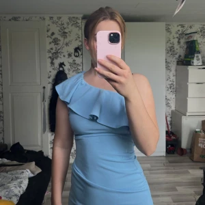 Ljusblå långklänning - Säljer en ljusblå långklänning med oneshoulder och snygg volangdetalj över axeln. Klänningen har en elegant och figurnära passform.  Aldrig använd 
