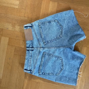 Blå jeansshorts med rå kant - Snygga blå jeansshorts med klassisk femficksdesign och rå, fransig kant nedtill. Shortsen har bälteshällor, knapp och dragkedja framtill samt stora bakfickor. Perfekta för en chill sommarlook. Använda 1-2 ggr. 