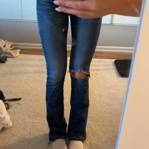 Mörkblå bootcut jeans med hål - Säljer ett par mörkblå jeans med bootcut passform och snyggt sliten look. Jeansen har ett stort hål på ena knät och klassiska fem fickor. Låg midja och stretchigt denimtyg som sitter skönt.