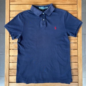 Ralph Lauren Piké Navy M - Säljer en klassisk marinblå pikétröja från Polo Ralph Lauren i slim fit. Tröjan har korta ärmar, krage med knappar och den ikoniska röda broderade loggan på bröstet. Tillverkad i bomull för en skön och stilren känsla.