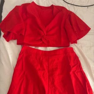 Röd croppad knytblus sett från SHEIN - Säljer en knallröd croppad blus från SHEIN med korta, vida ärmar och snygg knytning framtill. Toppen har v-ringning och är tillverkad i ett lätt, strukturerat tyg som känns luftigt och skönt. Perfekt för dig som vill sticka ut med färg! Med shorts till!