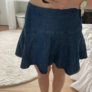Mörkblå jeanskjol med volangkant - Supersöt mörkblå jeanskjol i kort modell med volangig nederkant. Kjolen har en markerad midja och är sydd i klassiskt denimtyg. Perfekt för dig som gillar en lekfull och trendig stil. Passar till allt från sneakers till boots.