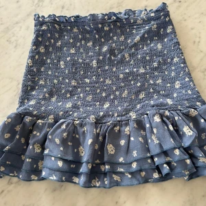 Blå blommig volangkjol - Supersöt blå kortkjol med vita blommor och smockad midja. Kjolen har flera lager volanger nertill och små volangdetaljer upptill. Perfekt för dig som gillar en feminin och trendig stil.