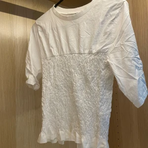 Trendig Vit smockad topp  - Supersöt trendig vit topp från H&M Divided med smockad midja och volangkant nertill. Toppen har rund halsringning och korta ärmar. 