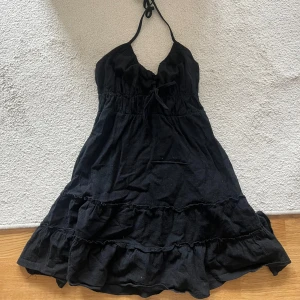 Svart halterneckklänning med volanger - Supersöt svart kortklänning med halterneck och knyt i ryggen. Klänningen har volangdetaljer nertill och en feminin v-ringning. Perfekt för varma dagar när du vill känna dig både chill och snygg.