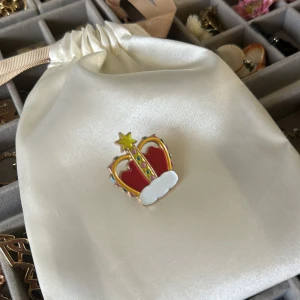 Färgglad krona brosch i emalj - Säljer en cool brosch i form av en krona med röd, gul och vit emalj samt små färgdetaljer. Kronan har en stjärna på toppen och fästs med nål på baksidan. Perfekt accessoar för att ge din outfit en kunglig touch.