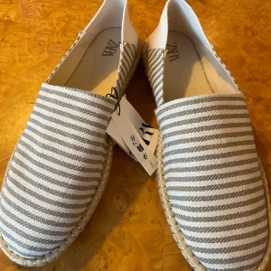 Oanvända espadrillos från Zara, strl 42 - Helt nya, oanvända espadrillos från Zara i storlek 42. Skorna har ett randigt mönster i ljusbeige/khaki och är tillverkade i bomullstyg med klassisk flätad sula. Perfekta för sommaren!