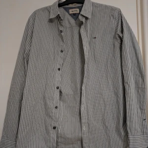 Randig skjorta från Tommy Hilfiger - Klassisk skjorta från Tommy Hilfiger med tunna vertikala ränder. Skjortan har lång ärm, knappar framtill och broderad logga på bröstet. Tillverkad i 100% bomull och har en normal passform. Perfekt för en clean och stilren look.