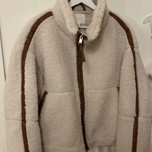 Beige teddyjacka med bruna detaljer - Mysig beige teddyjacka med bruna detaljer och dragkedja framtill. Perfekt för kyliga dagar med sin fluffiga och varma design. Jackan har långa ärmar och en hög krage för extra värme.