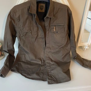 Brun skjorta från Northern Hunting - Säljer en brun skjorta från Northern Hunting i storlek M. Skjortan har långa ärmar och knappar framtill. Den har två bröstfickor, varav en med dragkedja. Perfekt för en avslappnad stil.är liten missfärgning på ryggen,man le se på bild , men man märker inte av det.kan möta er i Stockholm utanför Åhléns 