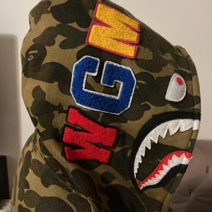 Hoodie från bape  - Säljer en bape Hoodie tror den heter så. med unika detaljer som hajtänder och broderade bokstäver på huvan. Hoodien har en dragkedja och är långärmad, perfekt för streetwear-stilen. Obs! Dragkedjan är jätte seg funkar inte att stänga!!!😊😊