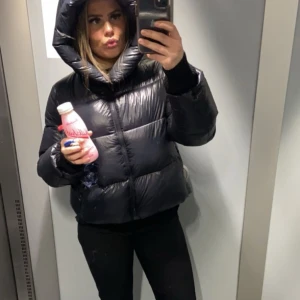 Svart pufferjacka - Säljer en snygg svart pufferjacka med huva. Jackan är perfekt för kyligare dagar och har en modern look. Den är kort och har en glansig yta. Passar perfekt för dig som vill hålla värmen med stil. Köpes för 2000 men pris kan diskuteras 