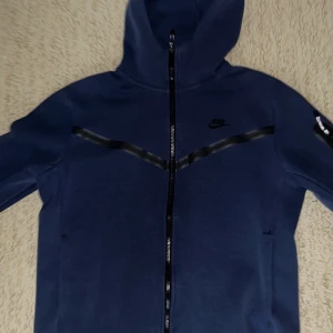 Mörkblå huvtröja från Nike - Säljer en mörkblå huvtröja från Nike med dragkedja och huva. Tröjan har långa ärmar och en diskret Nike-logga på bröstet. Perfekt för en sportig look eller avslappnade dagar. Den har även en praktisk ficka på ärmen.Den har en liten hål men man kan sy enkelt det är ingenting att oroa sig för 