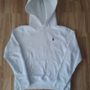 Vit hoodie från Polo Ralph Lauren - Säljer en stilren vit hoodie från Polo Ralph Lauren med det klassiska logotypbroderiet på bröstet. Tröjan har en känguruficka och ribbade muddar vid ärmslut och nederkant. Perfekt för en avslappnad look.