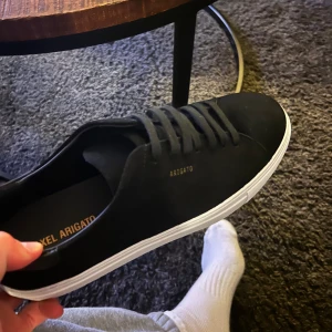 Svarta sneakers från Axel Arigato - Snygga svarta sneakers från Axel Arigato med vit sula och diskret logga i guld på sidan. Skorna har en stilren design med snörning och är tillverkade i mocka för en lyxig känsla.