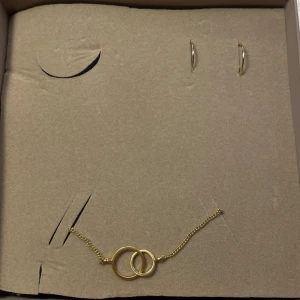 Guldhalsband och örhängen från Pilgrim - Helt nytt set med ett guldhalsband och matchande örhängen från Pilgrim. Halsbandet har en fin design med två sammanlänkade cirklar. Örhängena är enkla och stilrena, perfekta för att komplettera din look. Aldrig uttagna ur originalförpacknigen. Nypris 750